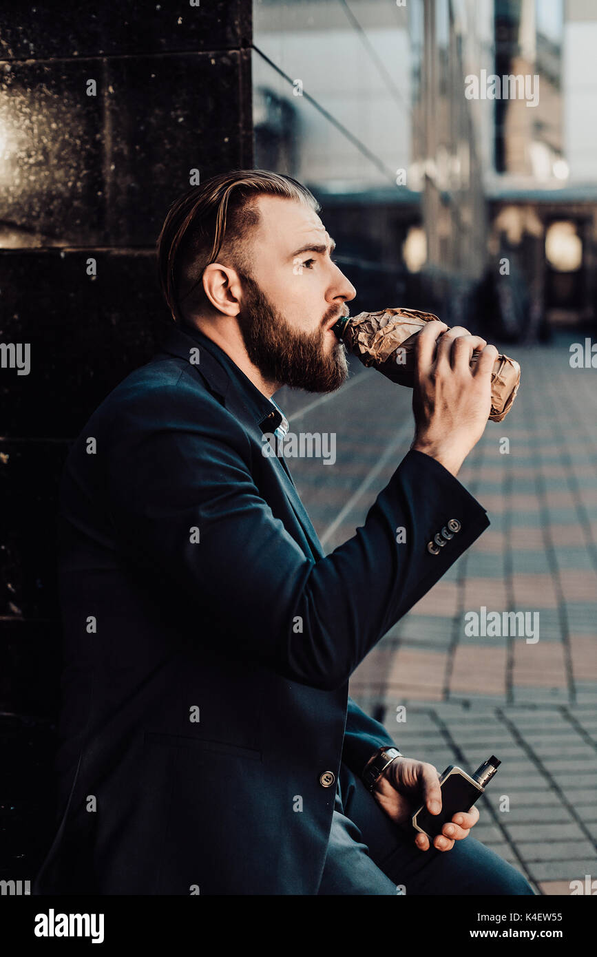 bearded man in a suit in the city drinking from a bottle of something Stock Photo