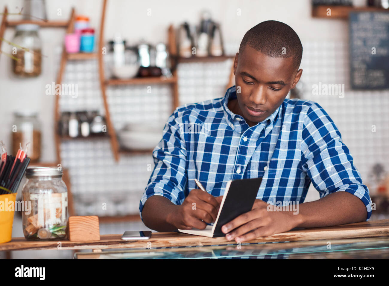 Focused young African entrepreneur standing at the counter of his trendy coffee shop writing down notes in a book Stock Photo