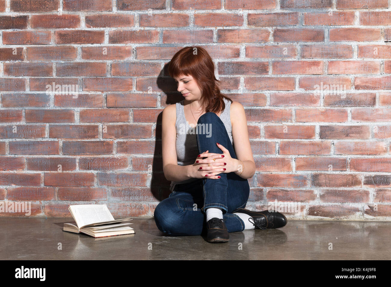 Young beautiful ginger hair woman in blue jeans reading a book while sitting on the floor near red brick wall. Stock Photo