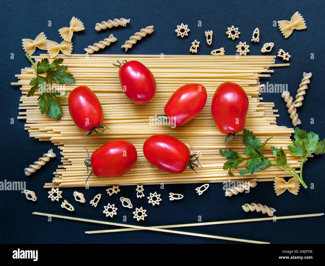 Spaghetti with tomatoes, greens and other figured pasta in the form of bows, spirals, figures of rockets and stars for cooking on a dark background Stock Photo