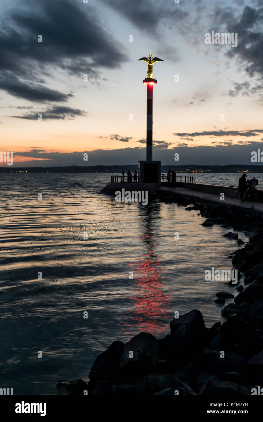 Lighthouse on lake at dusk.  Lighthouse, red light reflecting on calm water at dusk, dark clouds. Stock Photo
