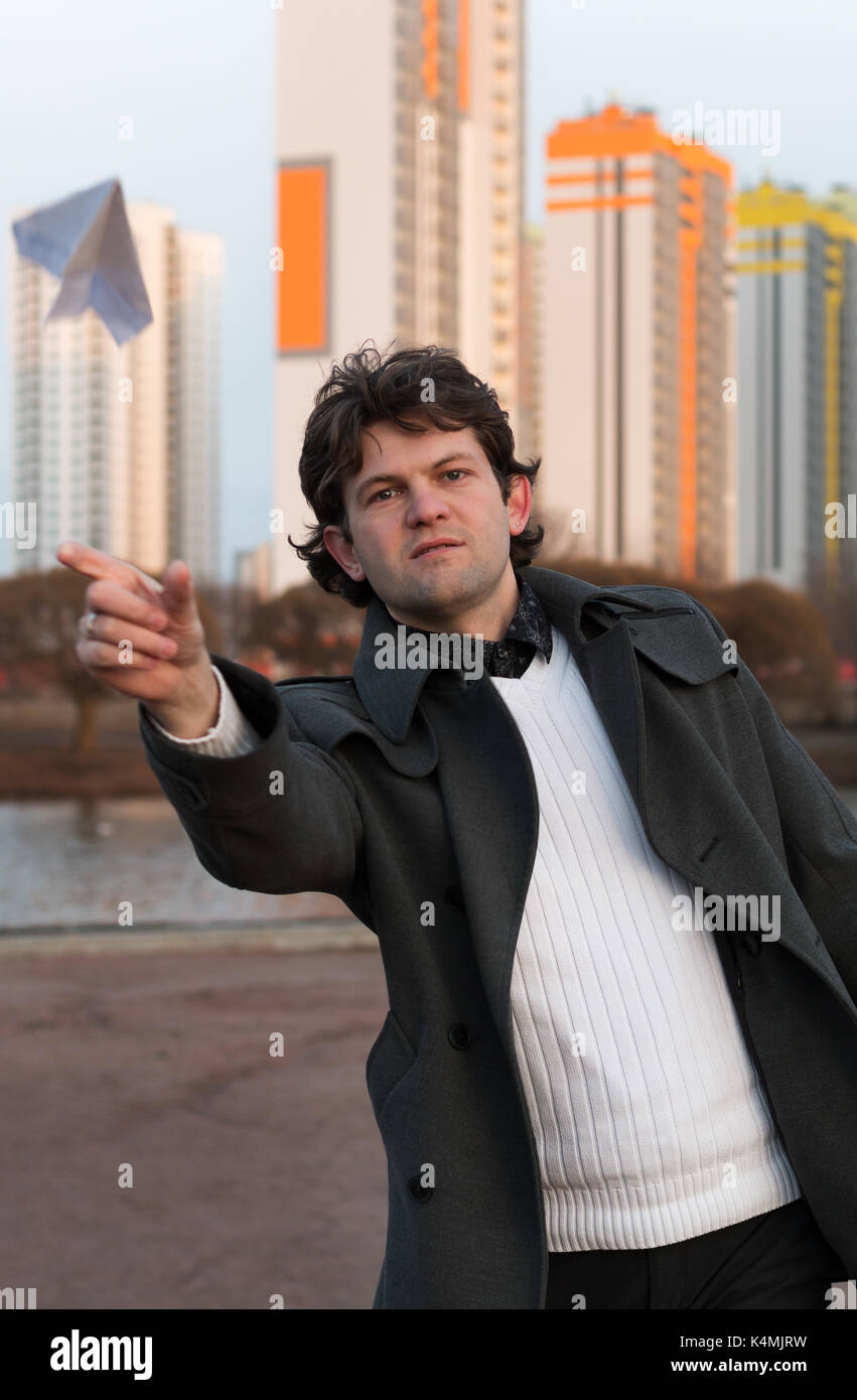 Paper airplane launching in park, free space, Man in grey coat and white sweater holding handmade paper airplane in hand on high-rise building background. Open-air game, rest, leisure, pastime concept Stock Photo