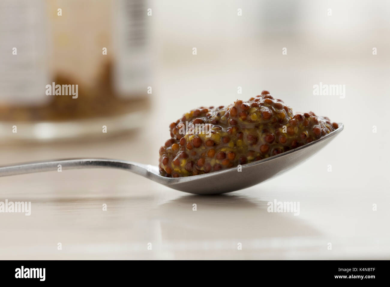 A heaped teaspoon of wholegrain mustard with the glass mustard jar forming part of the out of focus background. Shot in natural light. Stock Photo