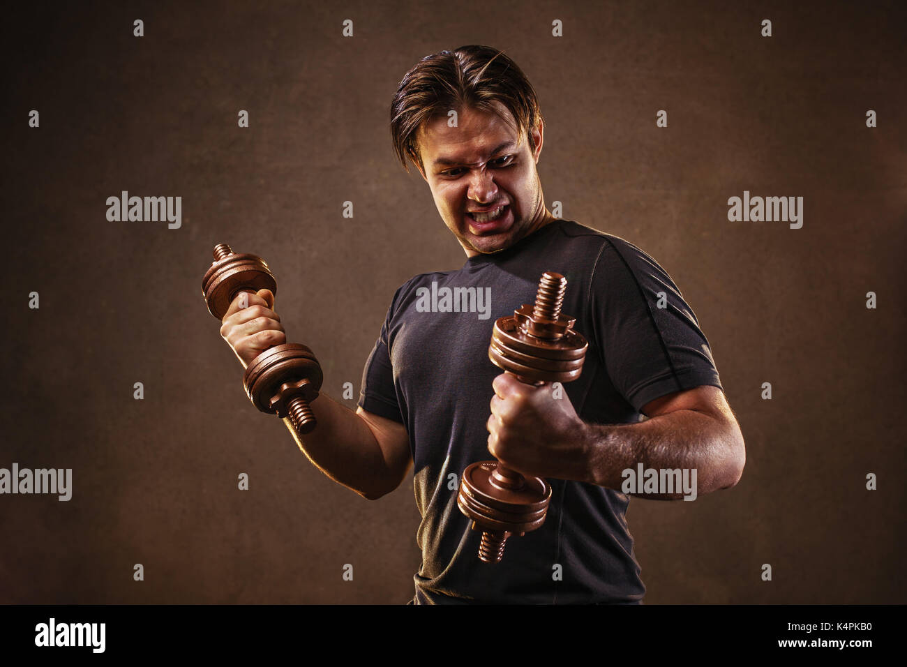 Young man hardly doing exercises with dumbbells on stone wall background. Stress and effort emotions. Stock Photo