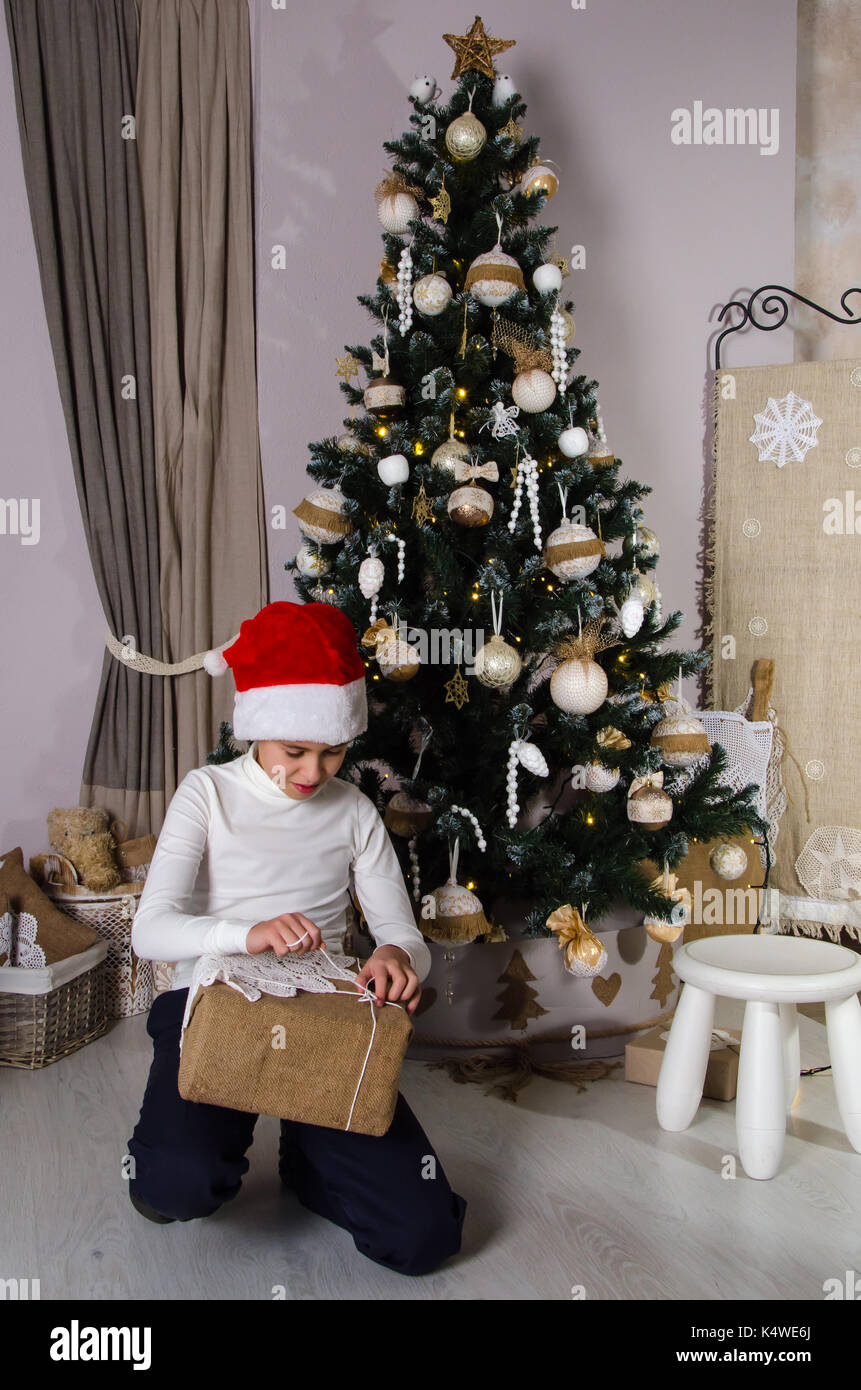 Boy wearing white sweater and Santa red hat opening Christmas gifts near decorated Christmas tree Stock Photo