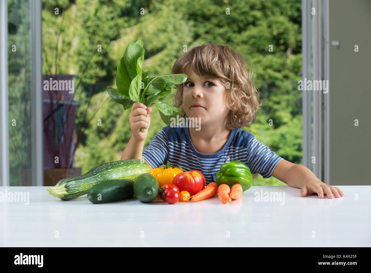 Cute little boy sitting at the table, expressing mixed emotions over vegetable meal, bad eating habits, nutrition and healthy eating concept Stock Photo