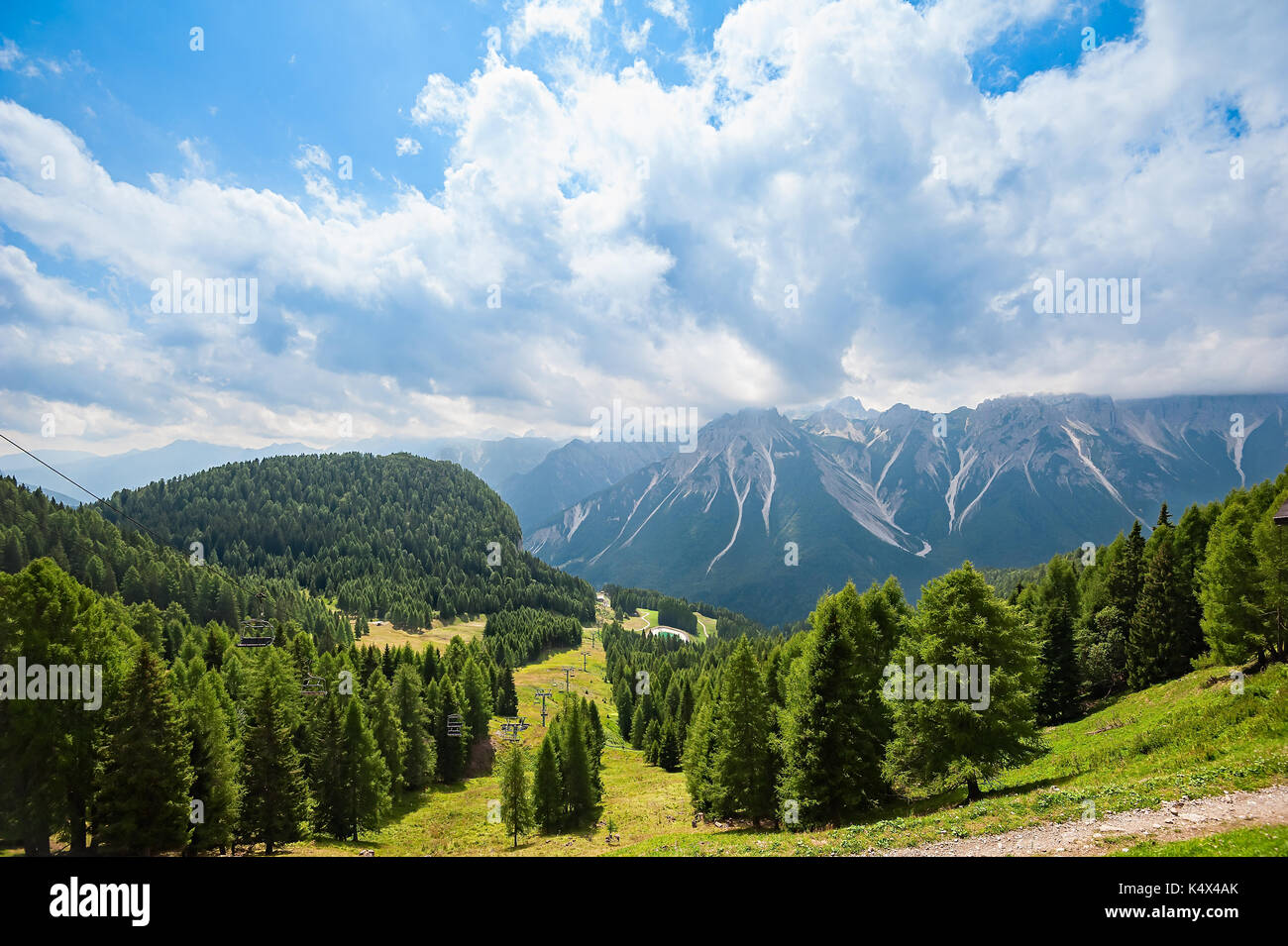Mountain panorama with forest, Dolomites, valley and chairlift in summer. Sky with clouds. Stock Photo