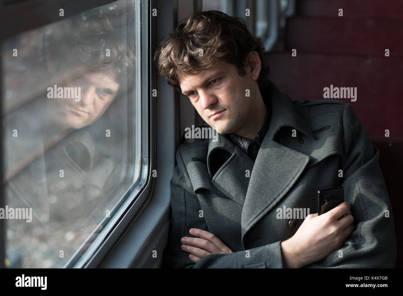 Traveling by train. Sad man traveling by train, looking through the window and thinking about unrequited love squeezing the phone in his hand. alone in an empty train wagon. Real people series. Stock Photo