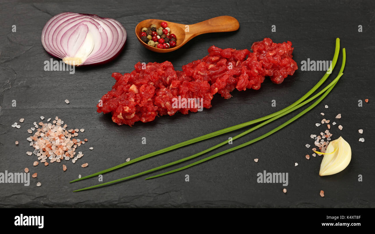 Raw minced beef red meat cutlet, spices, peppercorn in wooden scoop, spring green chive, onion, garlic cloves and Himalayan salt on black slate board, Stock Photo