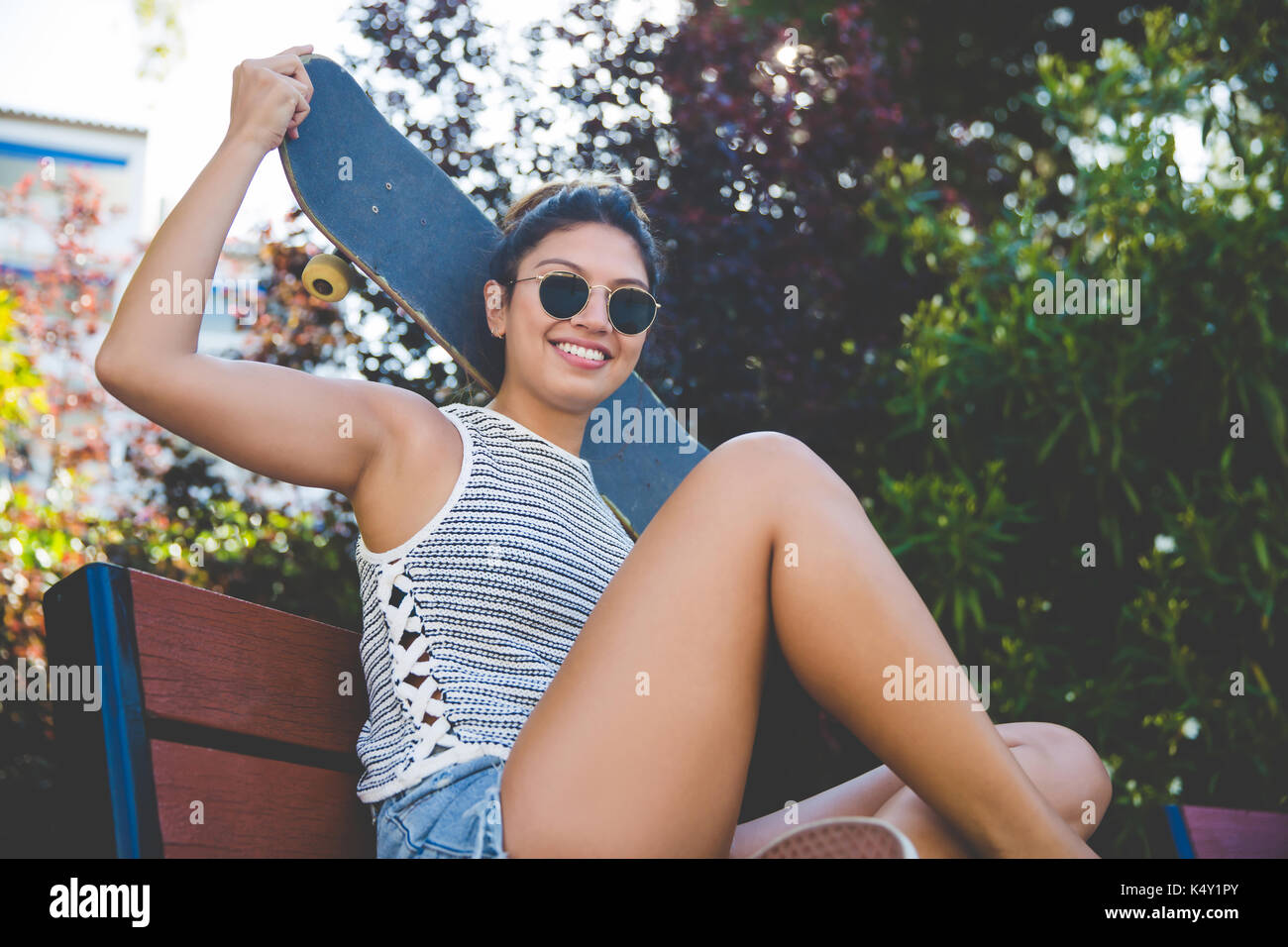 Portrait of young happy woman sitting on beach holding skateboard above head Stock Photo