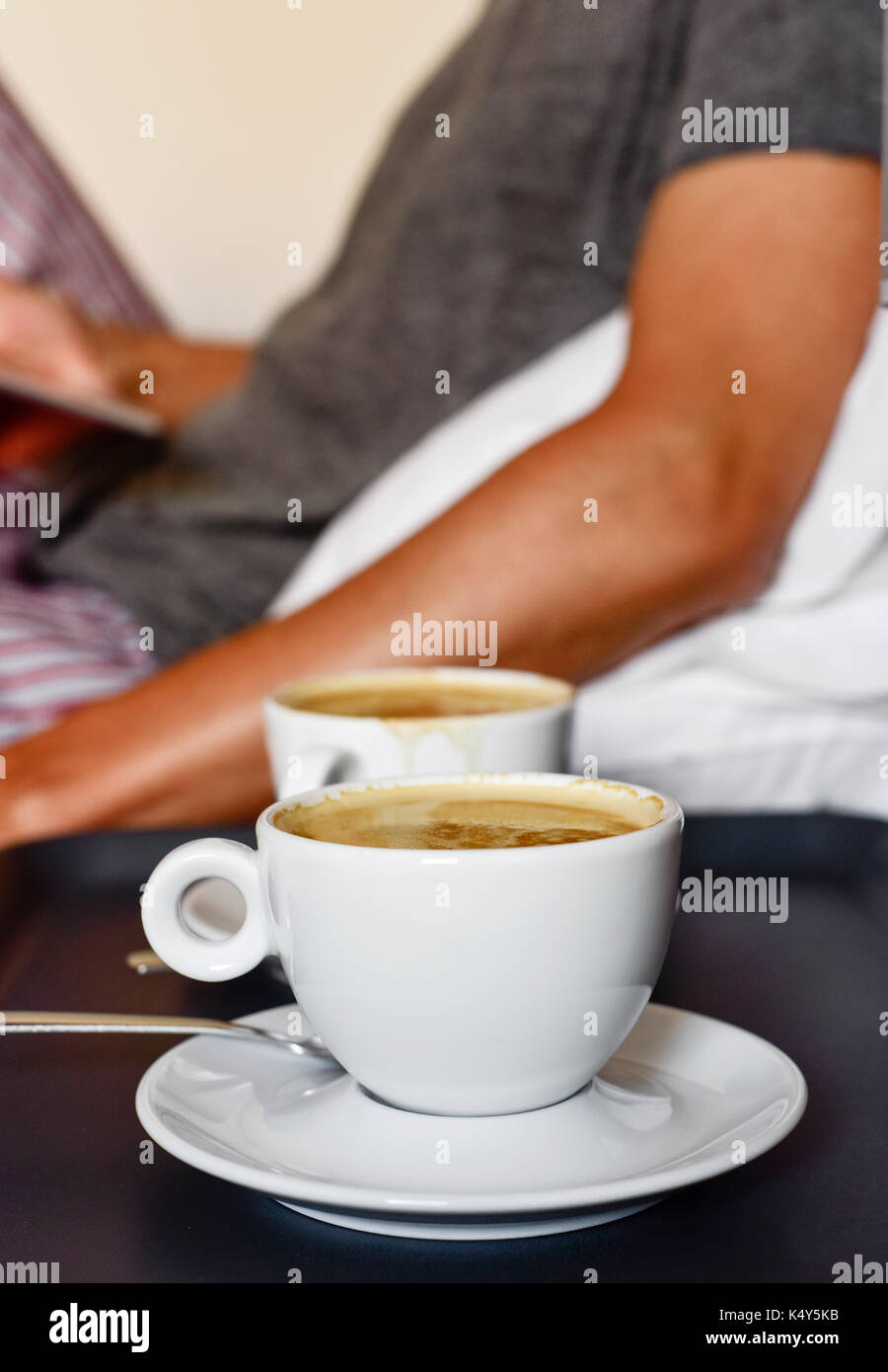 closeup of a pair of cups of coffee on a nightstand and a young caucasian man in pajamas in the background sitting in bed while is using a tablet or a Stock Photo