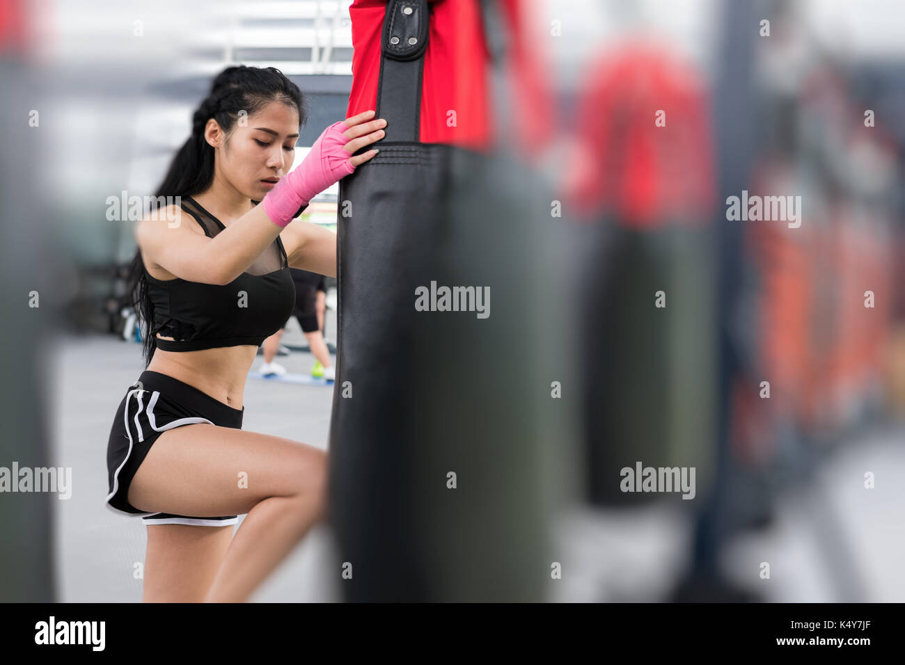 young woman execute exercise in fitness center. female athlete hitting and punching sandbag with rowing machine in boxing gym. sporty asian girl worki Stock Photo