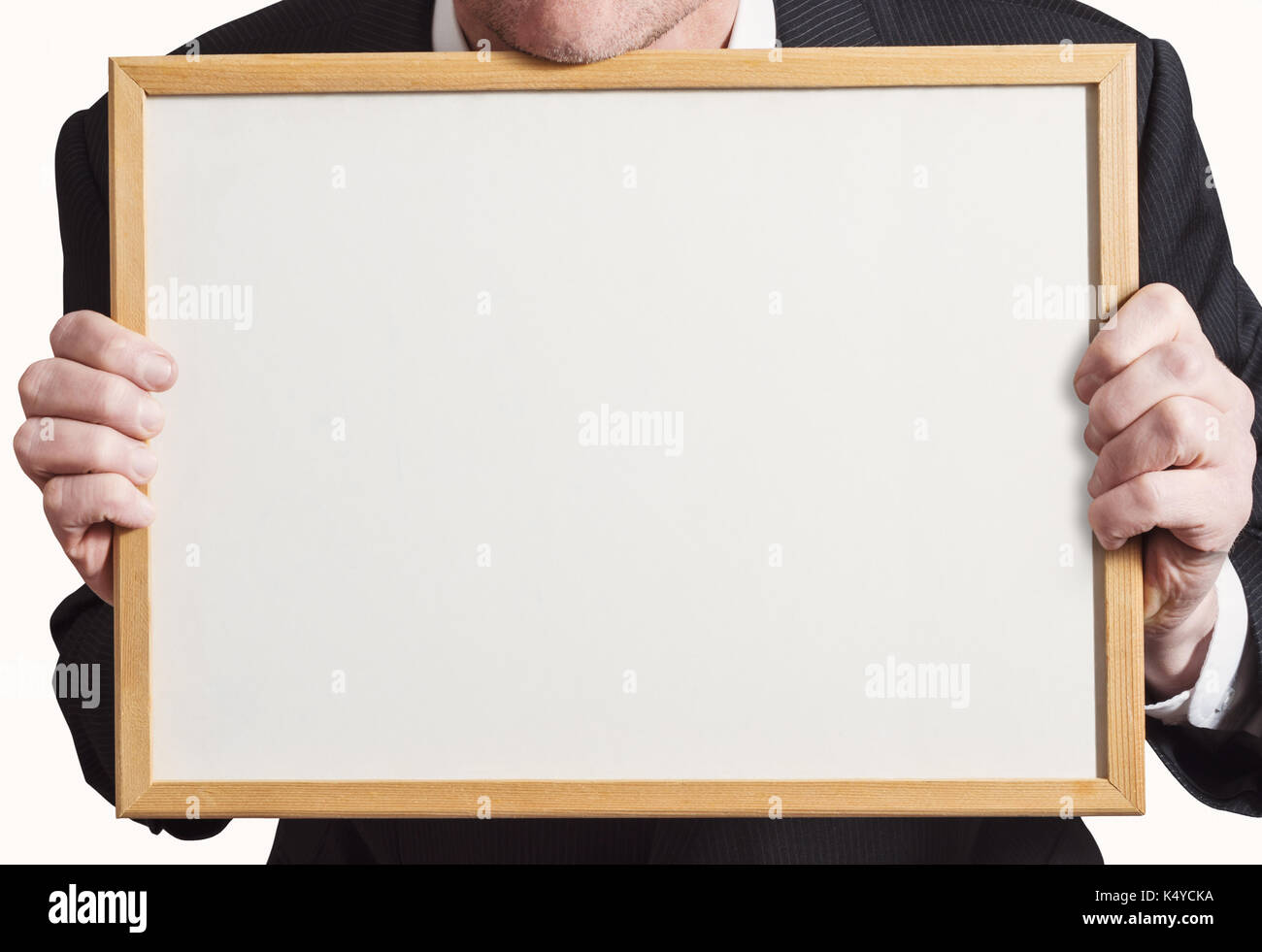 Unidentifiable businessman in dark suit, holding up a blank whiteboard with wood frame and facing front. Stock Photo