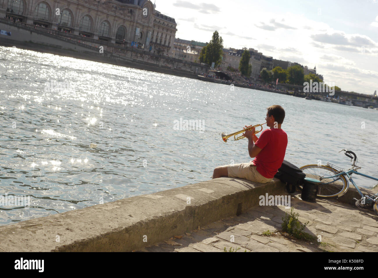 A man in a red t shirt practices the trumpet whilst sitting on the banks of the river Seine in Paris France.Summer time in the city. Stock Photo