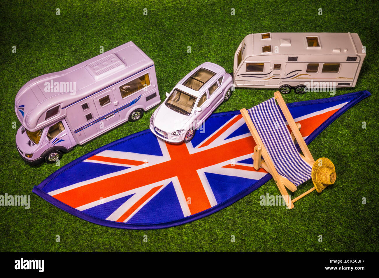 A model motorhome, car with caravan, deckchair and straw hat, on Union Jack colours. Concept of a UK motorhome or caravanning holiday or tour. Stock Photo