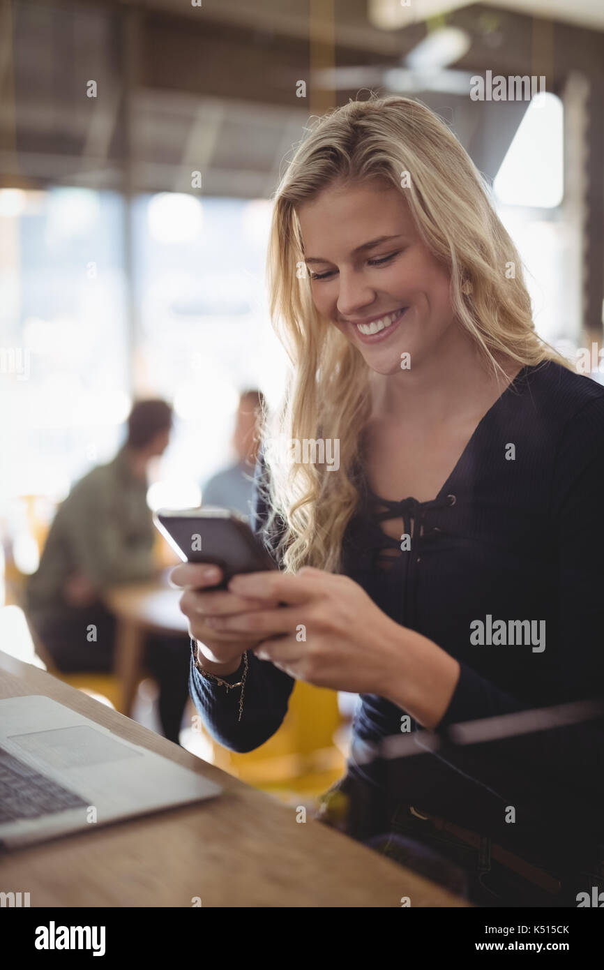 Smiling young blond woman texting through mobile phone while sitting at counter in coffee shop Stock Photo