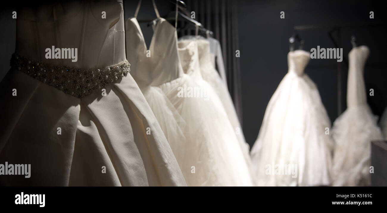 Selection of handmade white wedding gowns hanging on a rail in a darkened room partially illuminated by a light Stock Photo