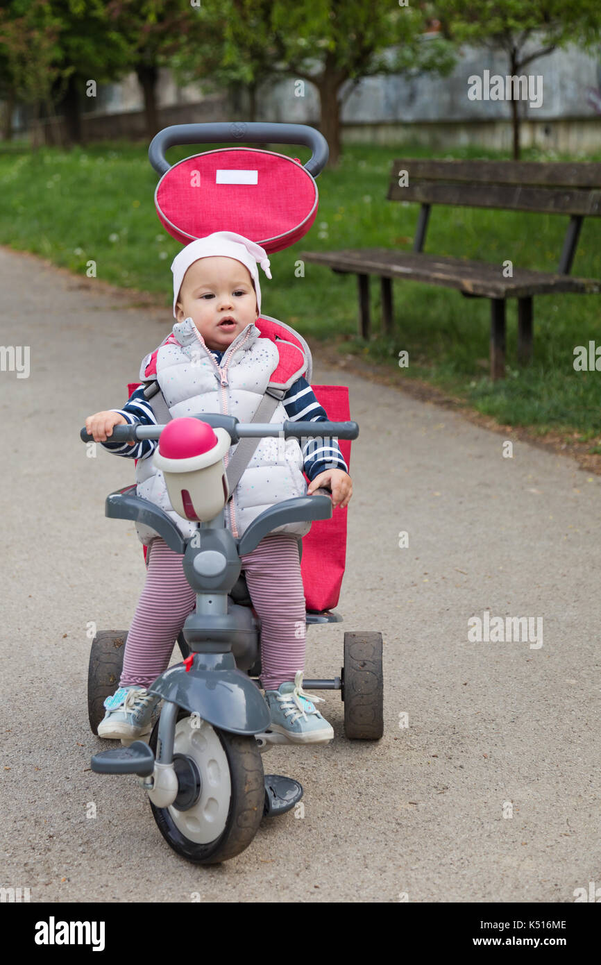 One year old baby girl sitting in a red and grey tricycle in the park by  the wooden bench Stock Photo - Alamy