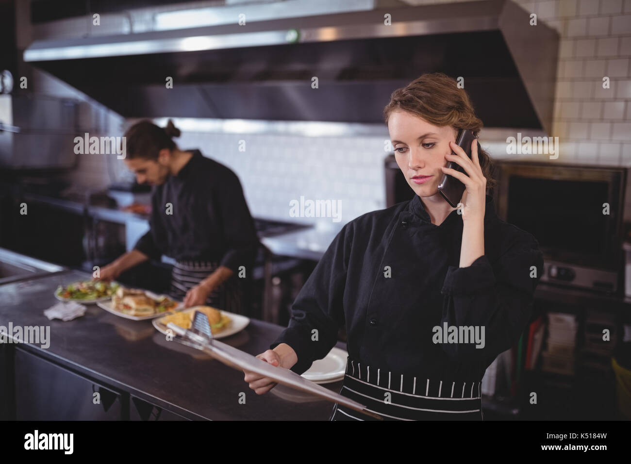 Young waitress talking on smartphone while waiter preparing food in
