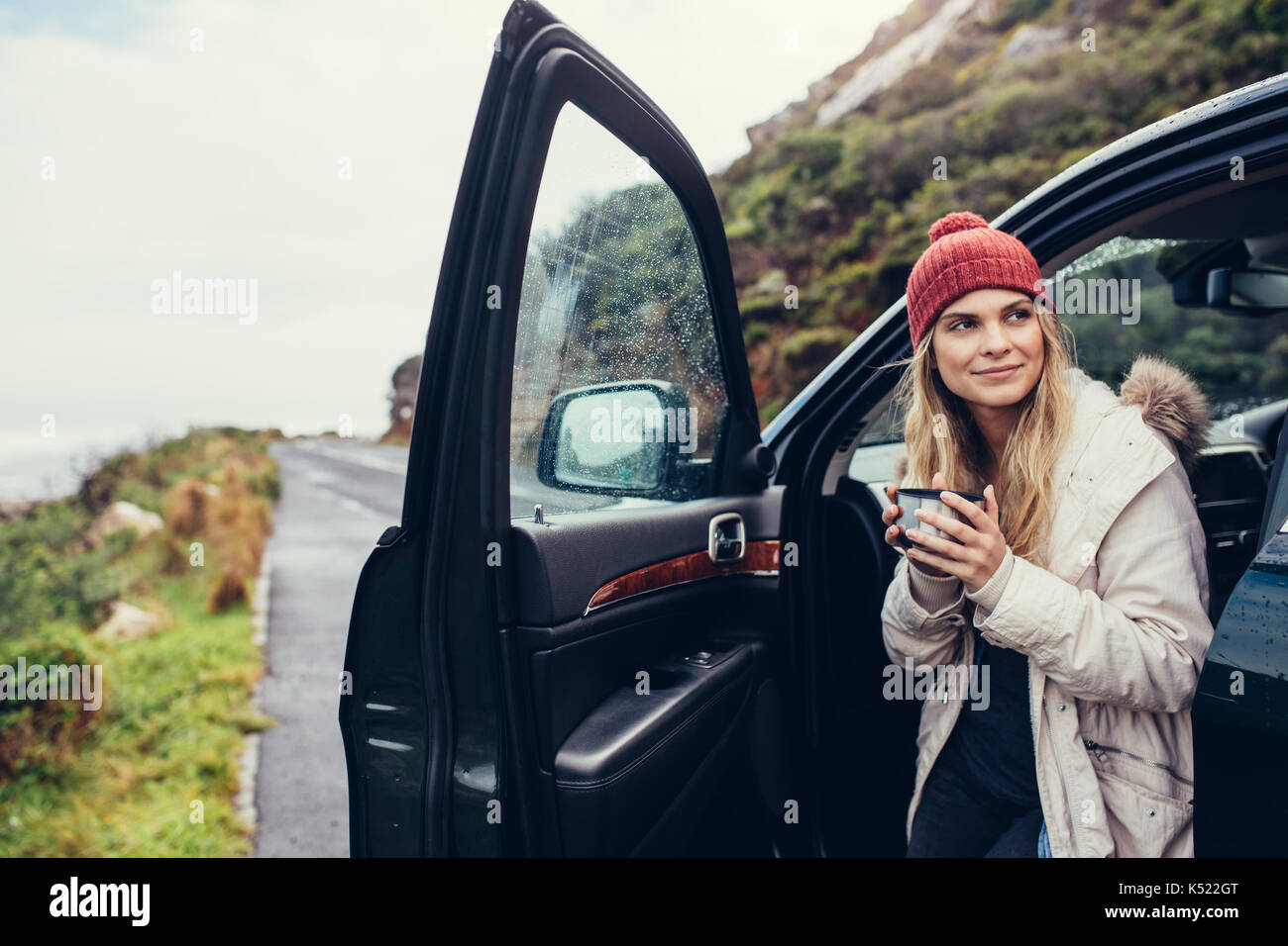 Beautiful female standing by the car with coffee. Smiling woman holding coffee and looking away. Stock Photo