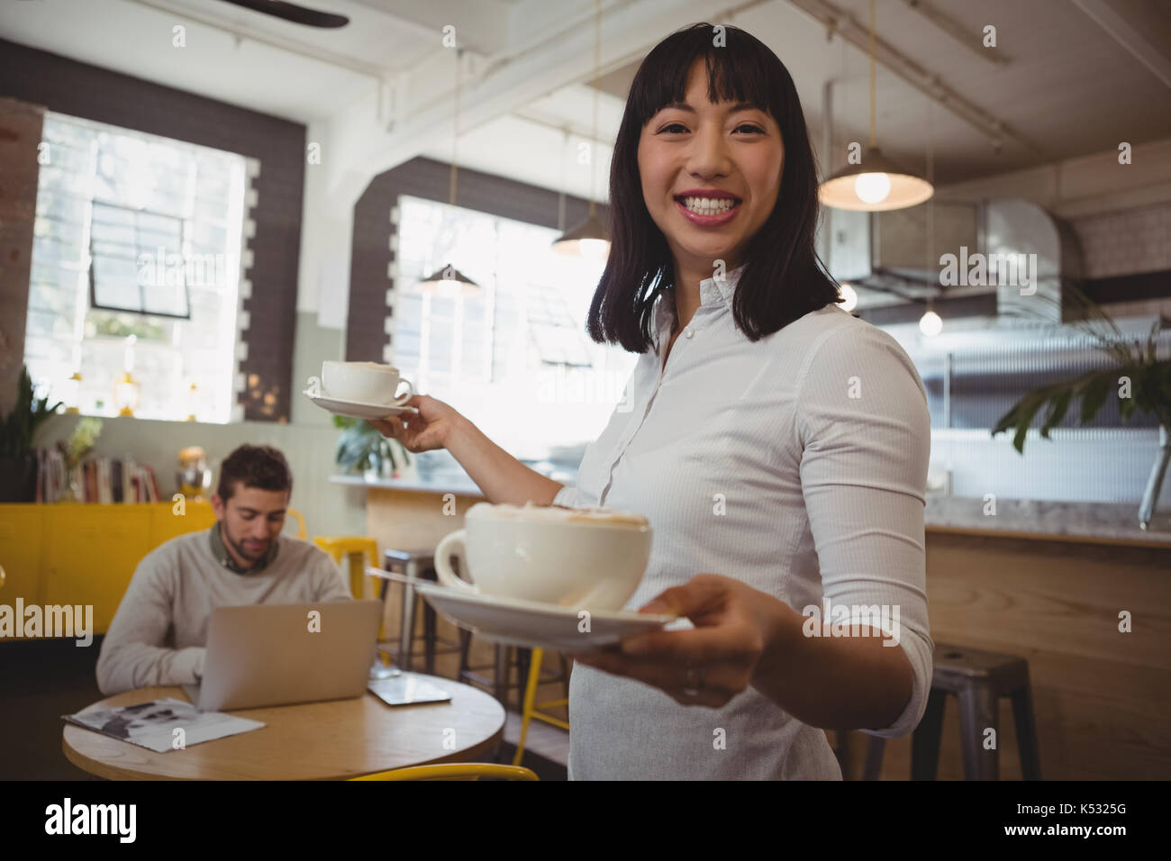 Portrait of smiling waitress holding coffee cups with man using laptop at table in cafe Stock Photo