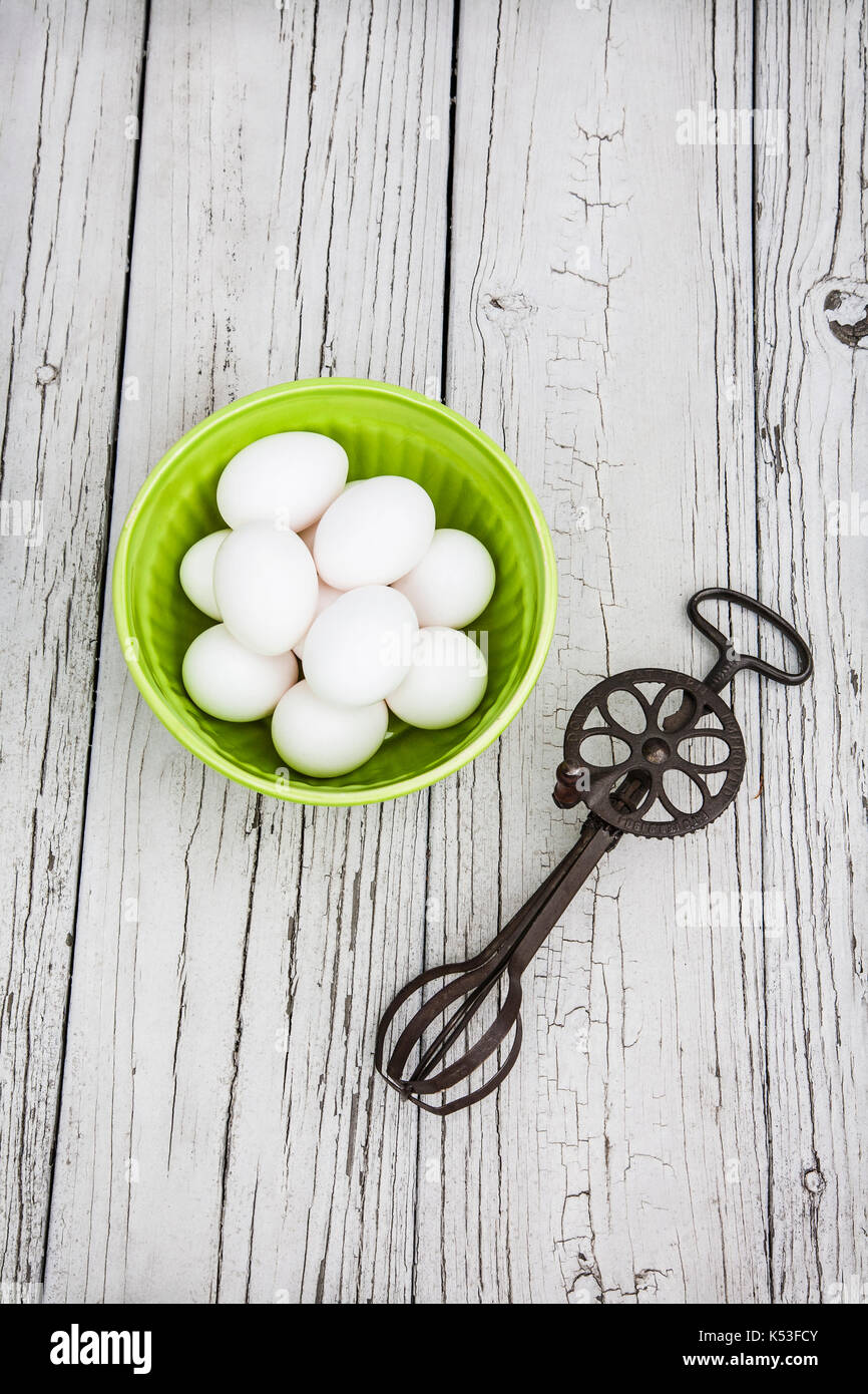 Cut out bowl of white eggs and an antique kitchen tool, eggbeater on weathered grey board backdrop, New Jersey, USA, hand tools, vintage baking tools Stock Photo