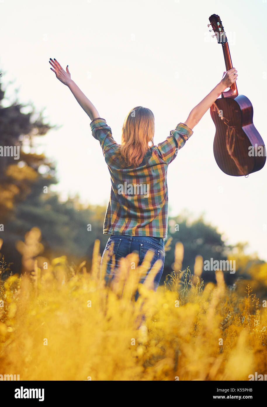 Photo from the back of a happy girl with her hands up with an acoustic guitar on a autumn meadow in sunset light. Stock Photo