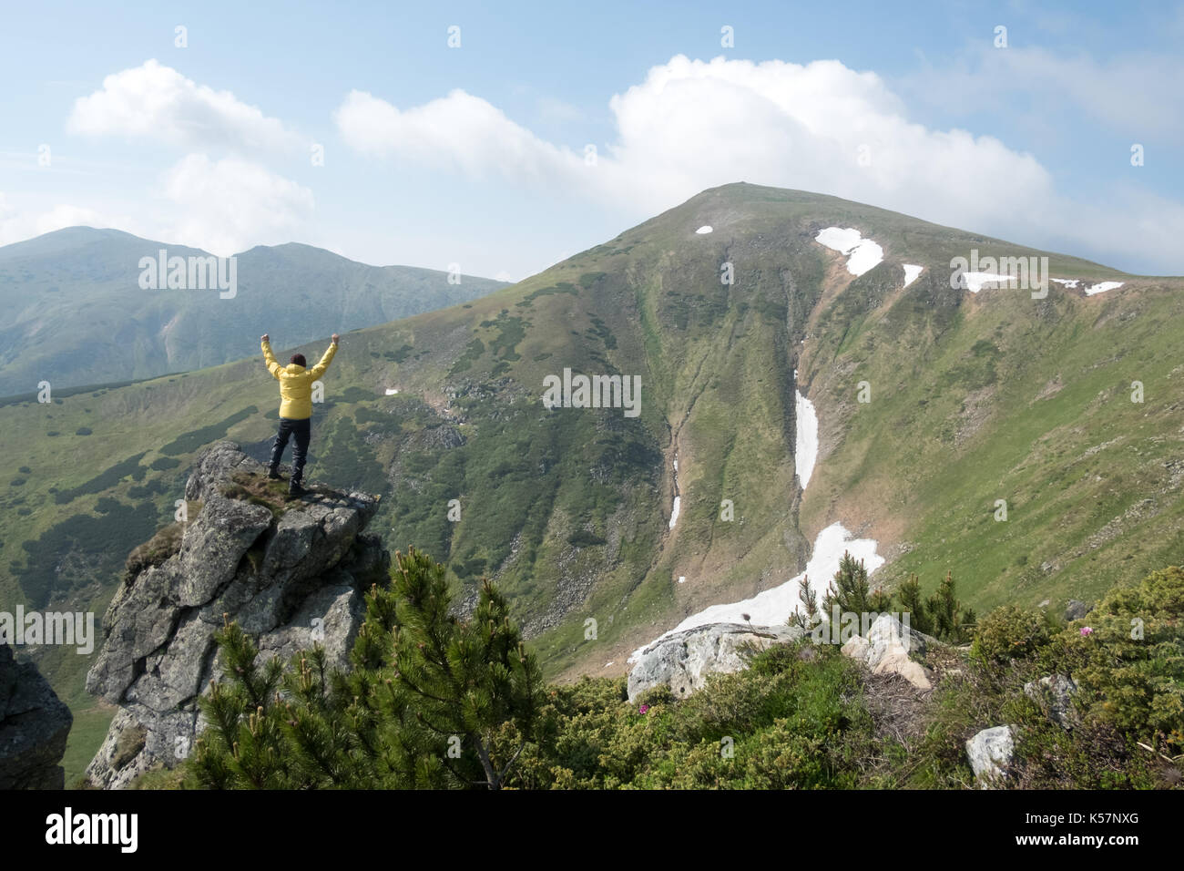 Alone tourist in yellow jacket stay on rock with hands up on high mountains. Stock Photo