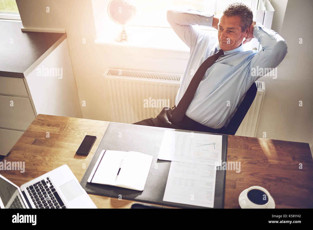 Smiling mature businessman in a shirt and tie leaning confidently back in his chair with his hands behind his head at his office desk Stock Photo