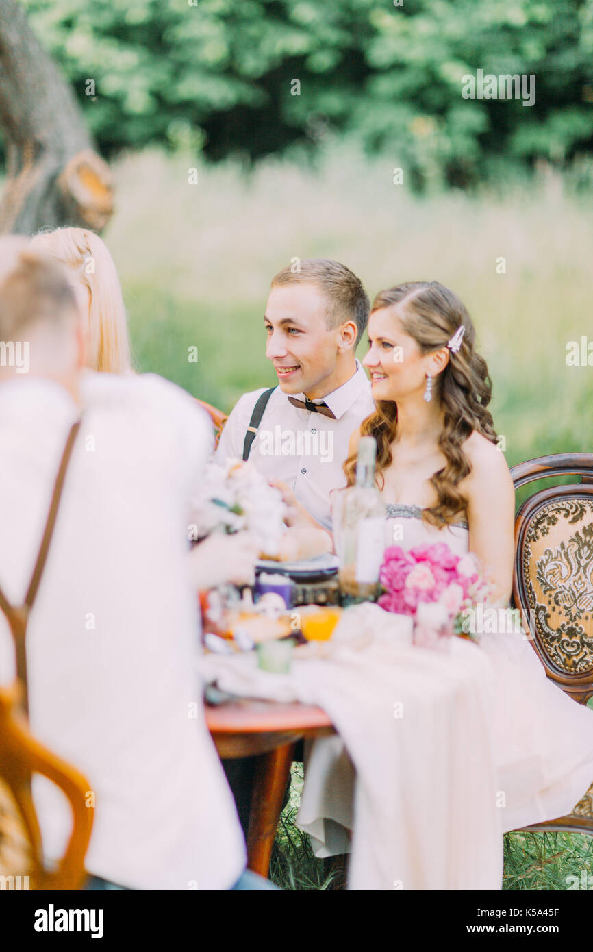 The side vertical photo of the bridesmaid in the long light pink dress and the best man with the wooden butterfly sitting at the wedding table. Stock Photo