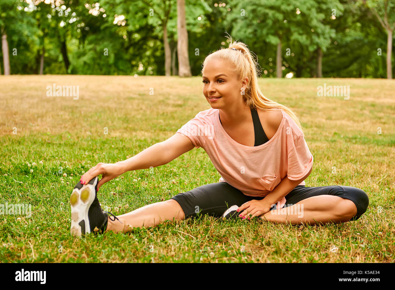A beautiful young blonde woman sitting in the grass and stretching her ...
