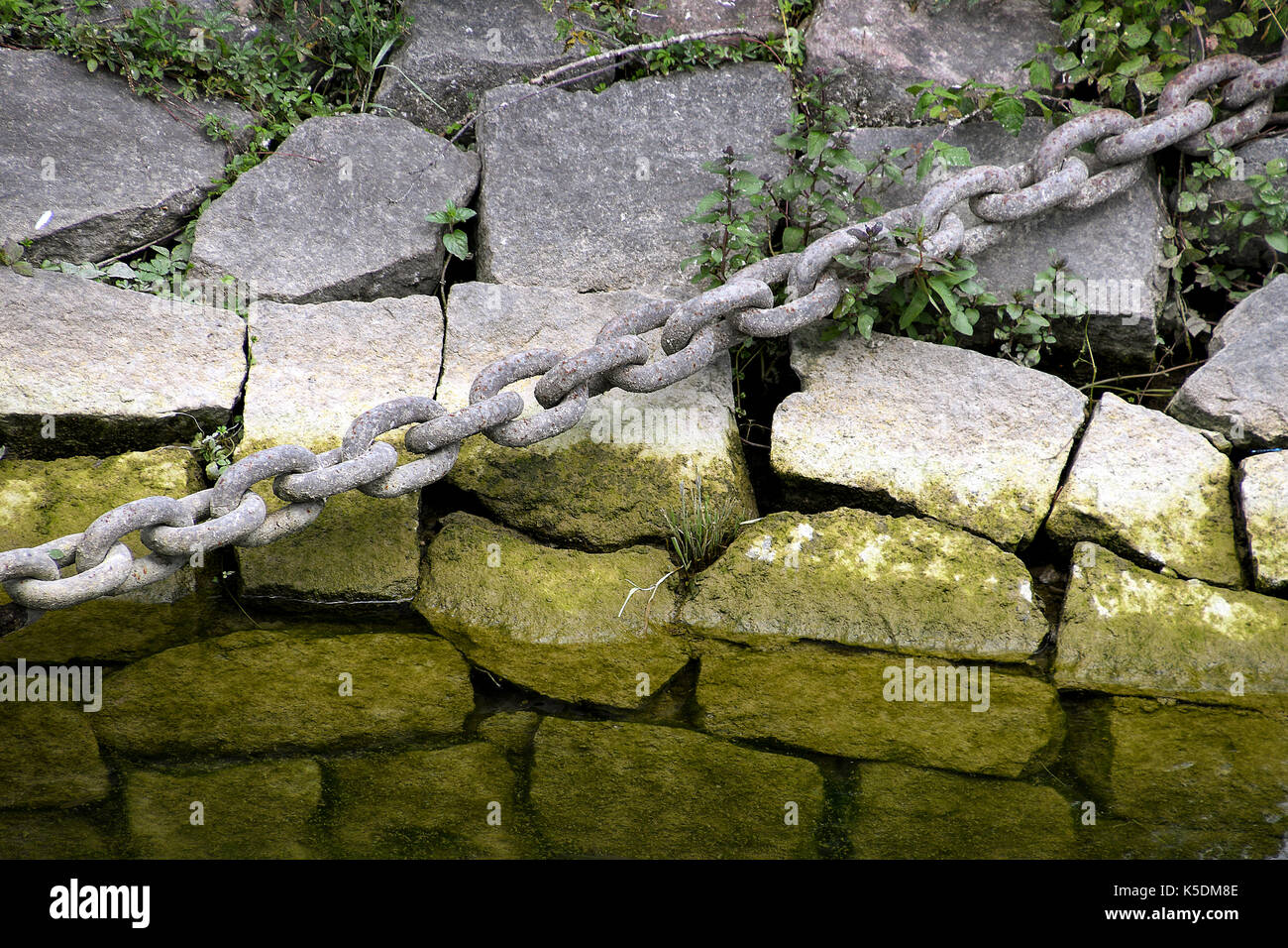 weathered iron chain over rocks with green algae Stock Photo