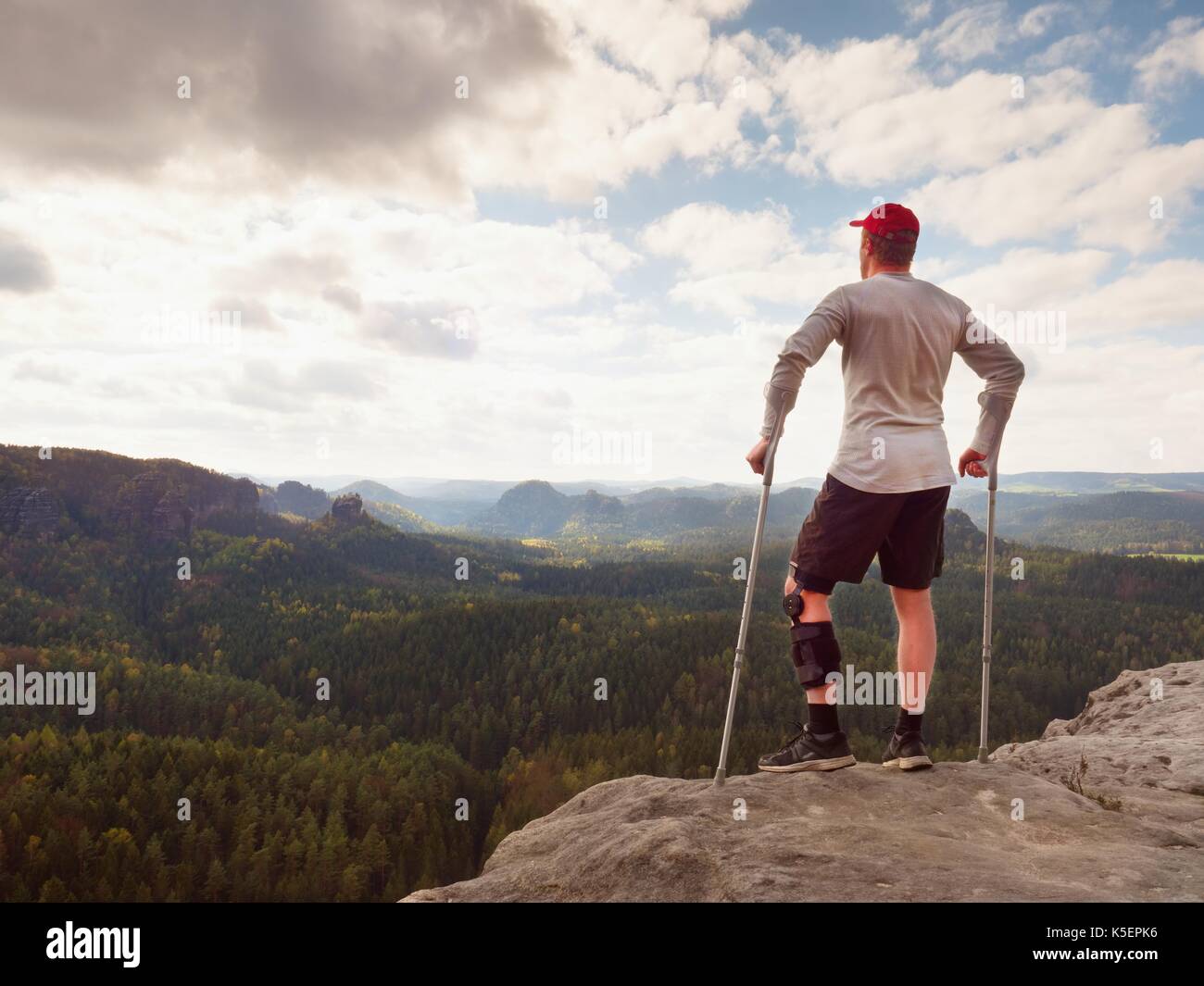 Hiker in grey t-shirt, medicine crutch and  leg fixed in immobilize achieved mountain peak. Man has fixed  broken leg in immobilizer Deep misty valley Stock Photo