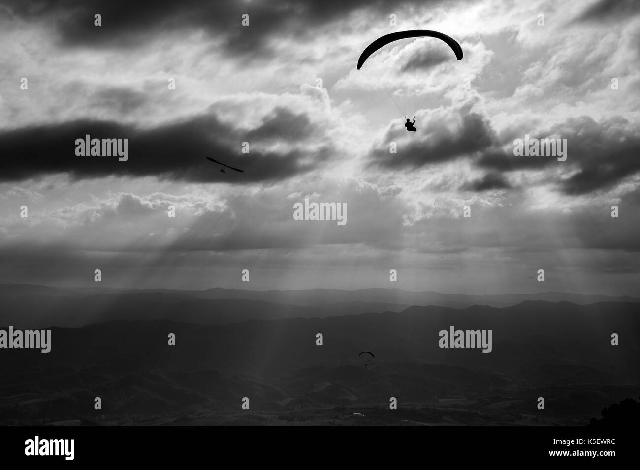 Some para-gliders and hang-gliders flying over a valley, with distant mountains and hills and sun rays coming out through the clouds Stock Photo