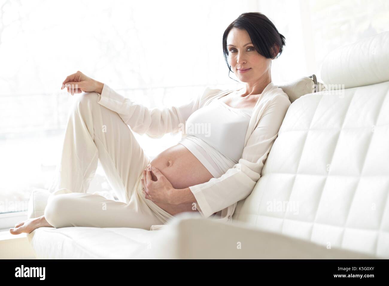 Pregnant woman sitting on sofa touching tummy. Stock Photo