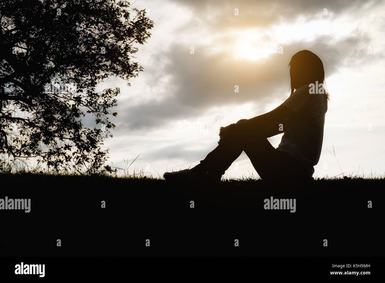Sad and depressed woman sitting alone at the field during beautiful sunset with park background. Selective focus. Stock Photo