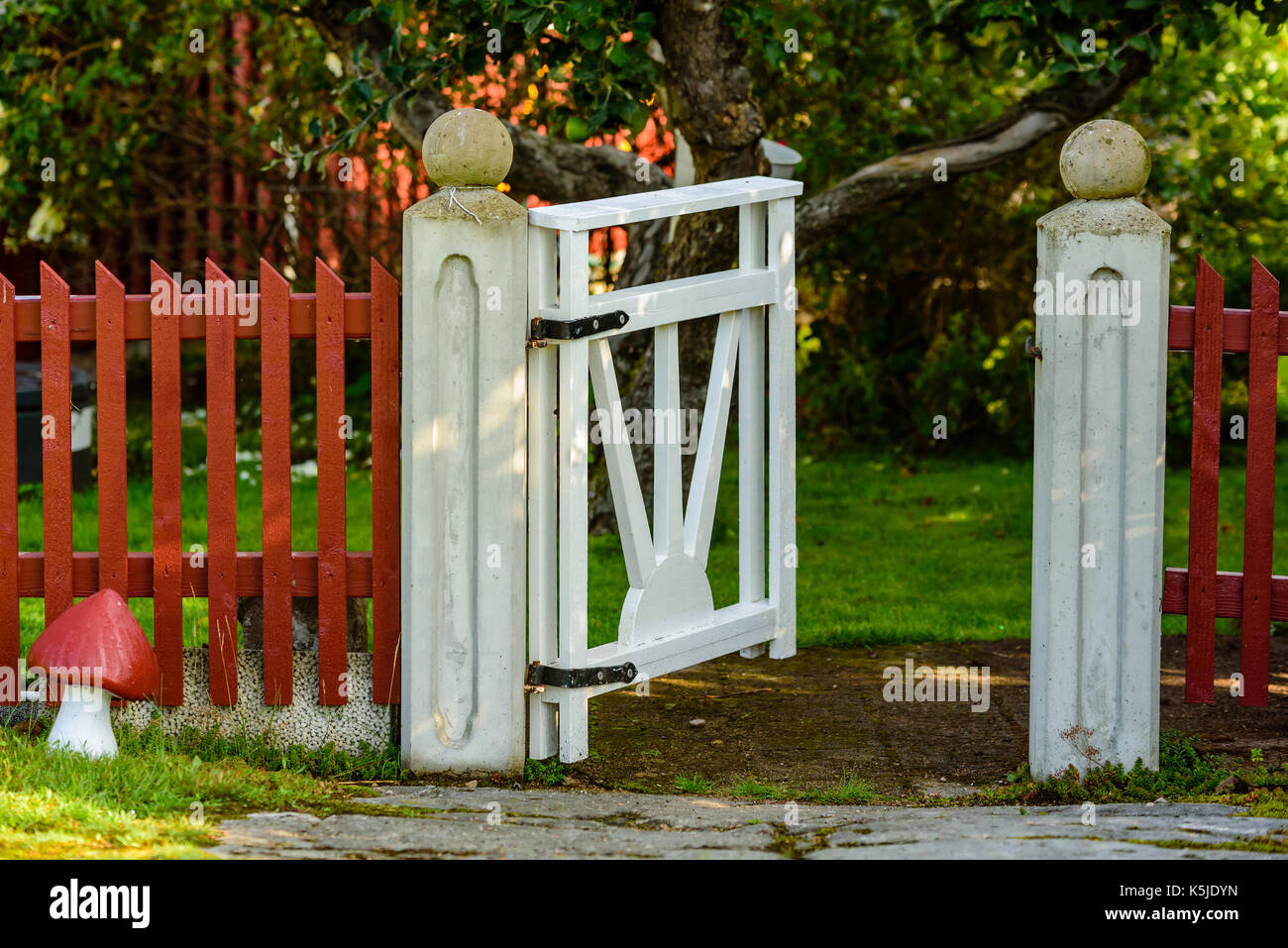 Open white garden gate on red picket fence. Lane and tree in background. Stock Photo