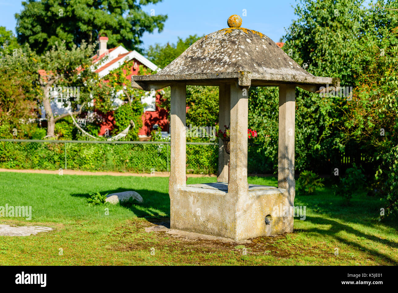 Public concrete water well with flower basket hanging inside it. Stock Photo