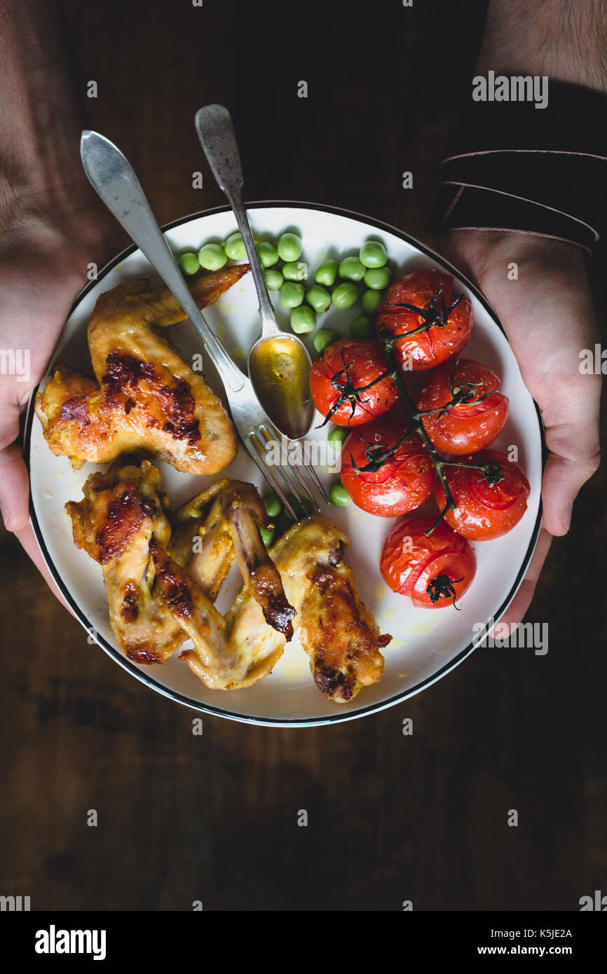 Plate with grilled spicy chicken wings, tomatoes and green peas in man's hands. Concept of balanced meal. Table top view, selective focus Stock Photo