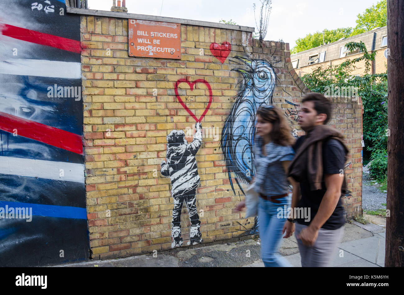 A couple walk past a brick wall decorated in street art featuring a pigeon and a boy drawing a red heart. Stock Photo