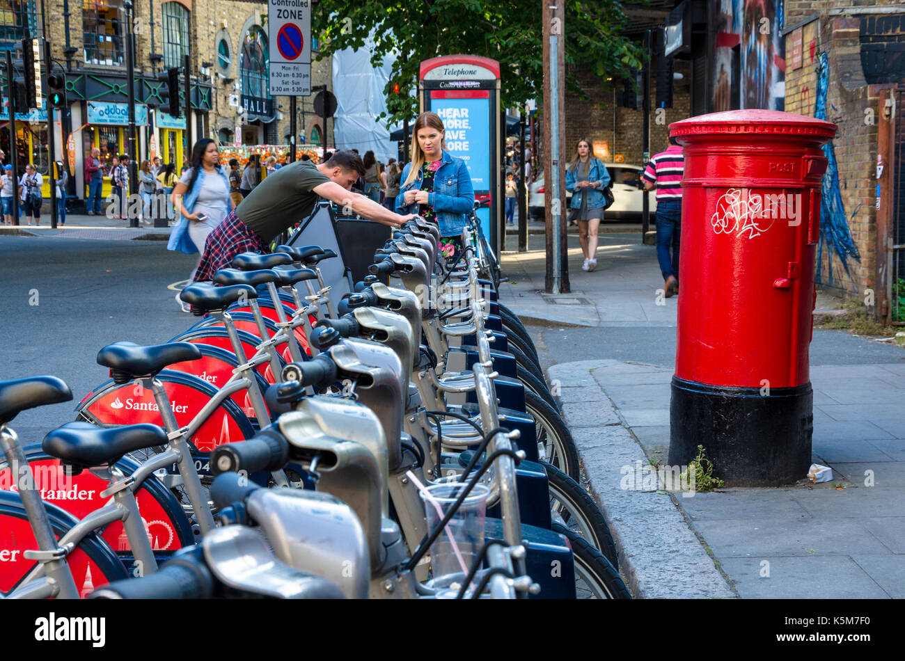 A man and woman try and work out how to hire a Santander bike form the docking station on Castlehaven Road in Camden Town, London. Stock Photo