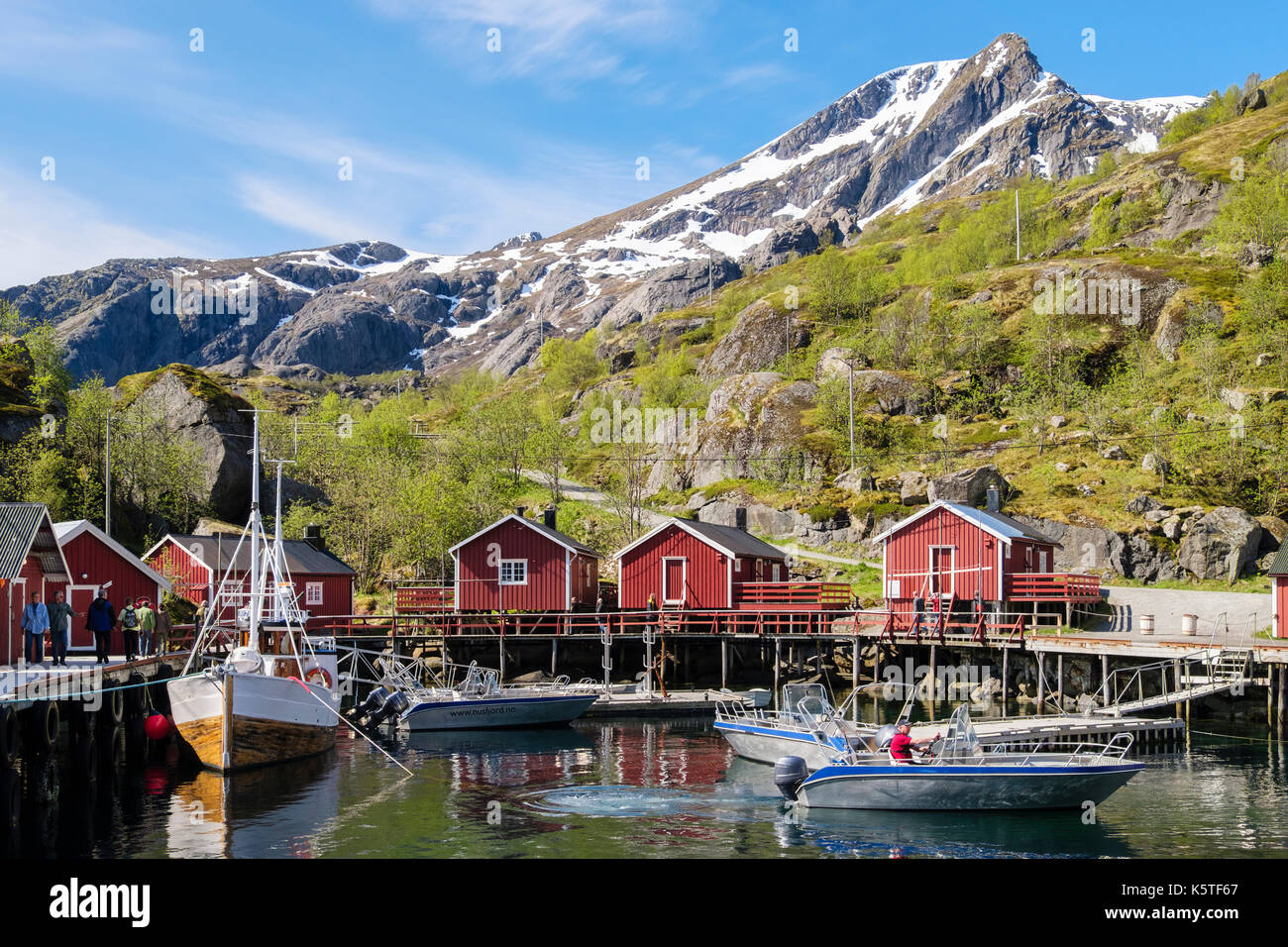 Boats Rorbu and tourists in harbour of historic fishing village in stunning landscape. Nusfjord, Flakstadøya Island, Lofoten Islands, Nordland, Norway Stock Photo