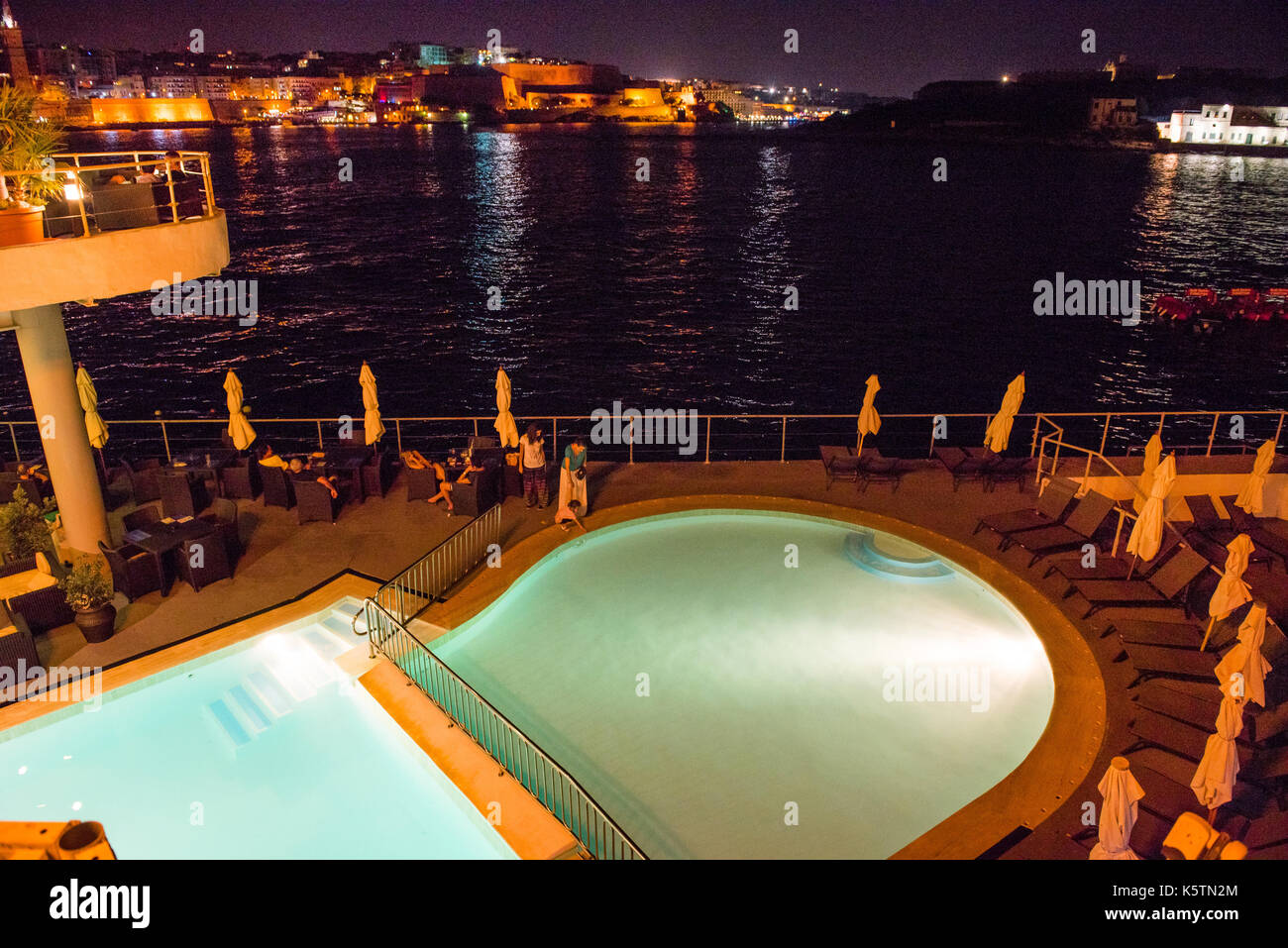 SLIEMA, MALTA - AUGUST 22, 2017: Tourists relaxing near a sea luxurios swimming pool at night with a great panoramic night cityscape to Valletta Stock Photo