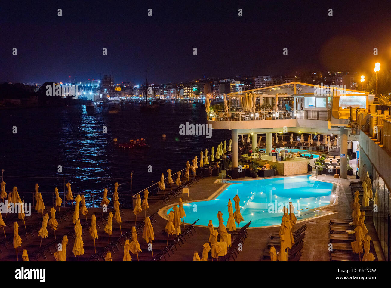 SLIEMA, MALTA - AUGUST 22, 2017: Tourists relaxing near a sea luxurios swimming pool at night with a great panoramic night cityscape to Valletta Stock Photo