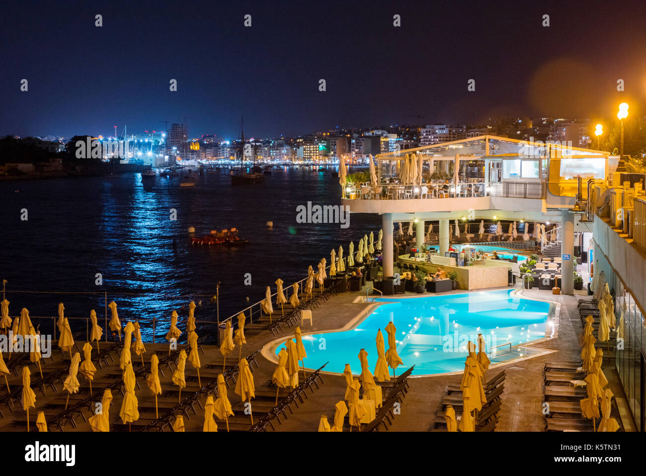 SLIEMA, MALTA - AUGUST 22, 2017: Tourists relaxing near a sea luxurios swimming pool at night with a great panoramic night cityscape to Valletta Stock Photo