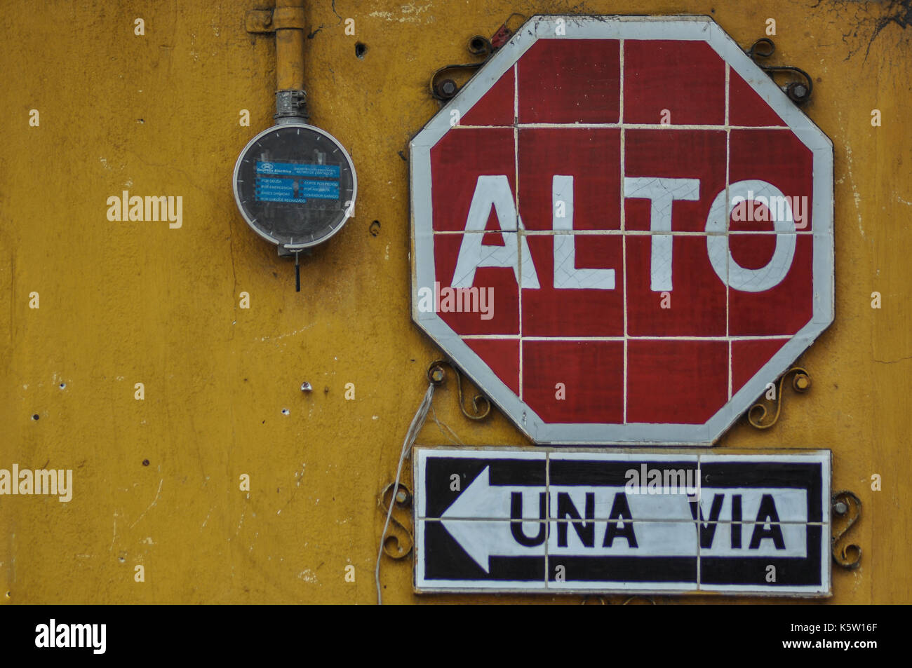 A red and white stop sign reading 'alto' is mounted on a yellow wall above a black and white sign indicating 'una via,' which translates to 'one way' Stock Photo