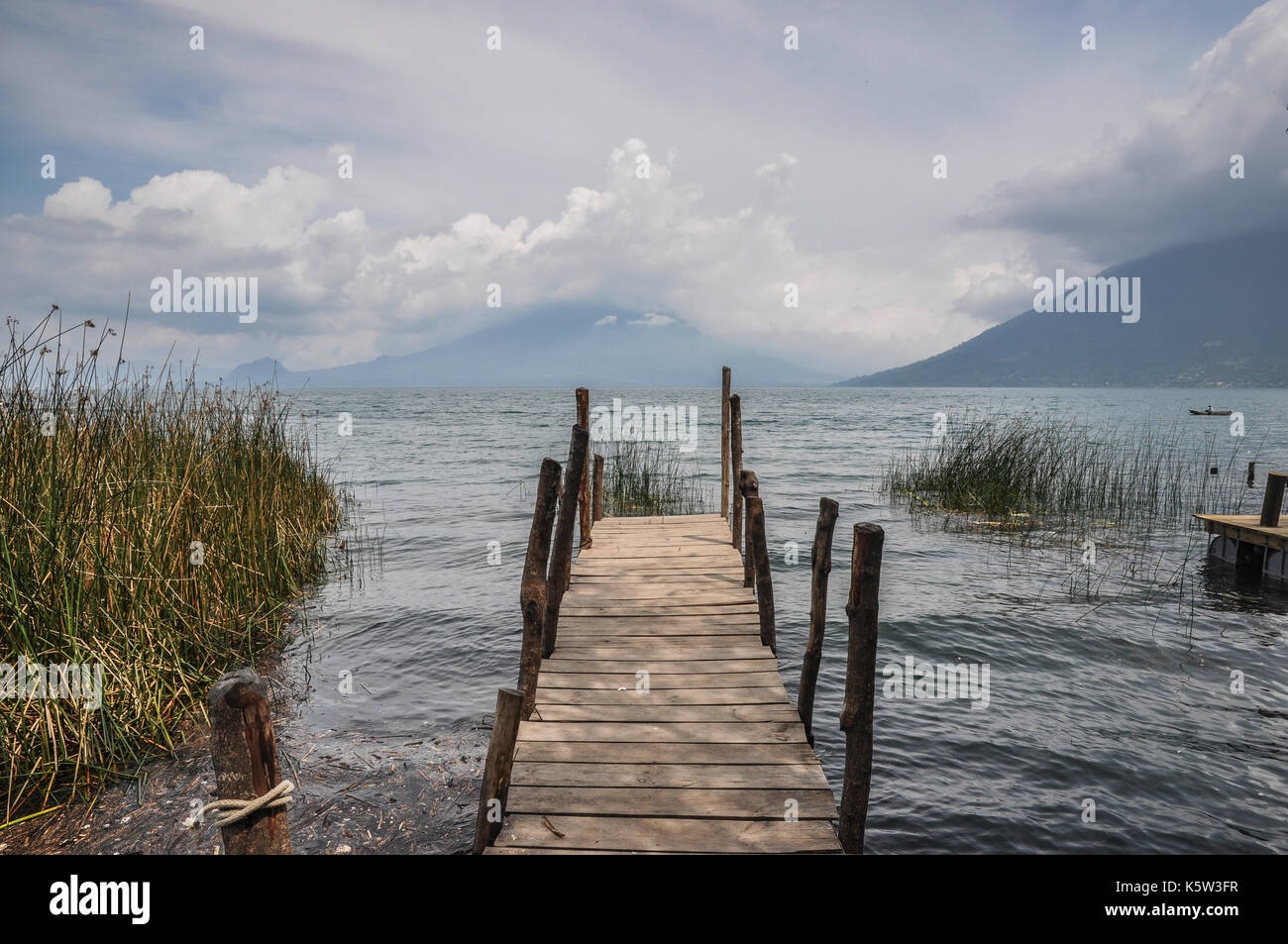 A wooden pier extends into a calm lake surrounded by mountains and under a cloudy sky. Stock Photo