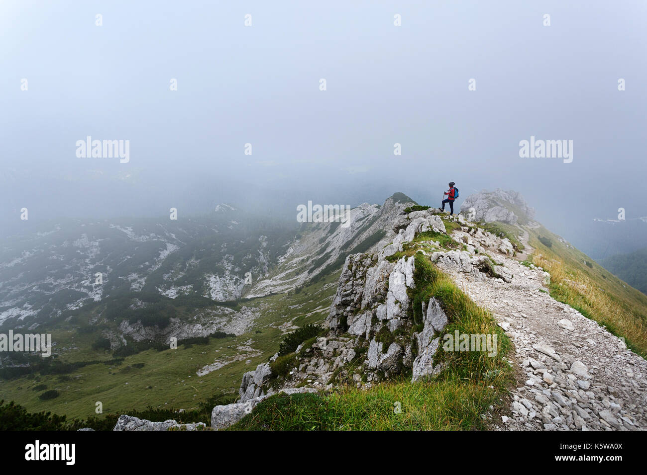 Woman in red windproof jacket standing on top of the mountain covered in fog, Visevnik, Julian Alps, Slovenia. Stock Photo