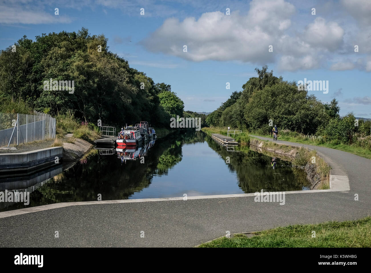 Canal boat moored at the jetty near the Falkirk Wheel Visitor's Centre as cyclists pass by - on the Scottish Canals in Falkirk, Scotland Stock Photo