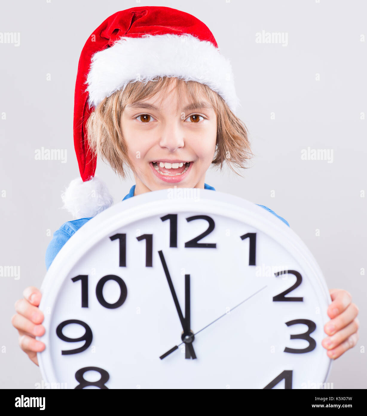 Emotional portrait of attractive caucasian little girl wearing Santa Claus red hat. Funny cute smiling child 10 year old with big clock on gray backgr Stock Photo
